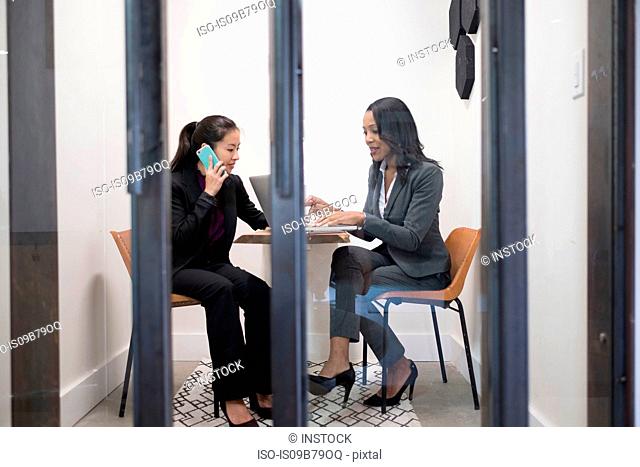Two businesswomen in office, using laptop, talking on smartphone