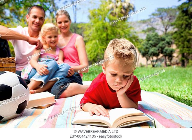 Concentrated blond boy reading while having a picnic with his family