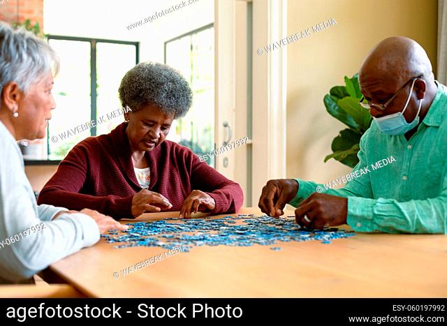 Two diverse senior women and african american male friend wearign face mask doing puzzles