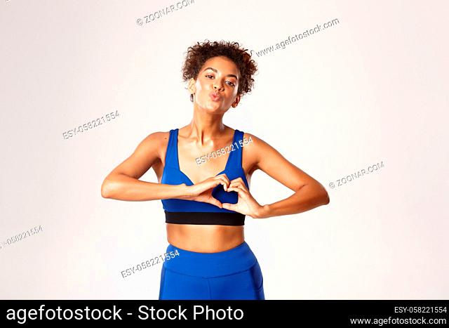 Concept of sport and workout.Beautiful young and healthy fitness woman in blue gym outfit, showing heart gesture and kissing someone, white background