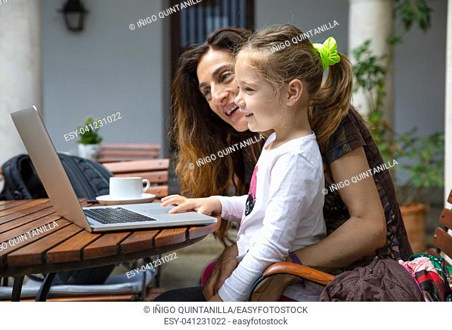 girl four years old sitting on mother legs, surfing internet and smiling watching laptop pc computer, with coffee cup on brown wooden table