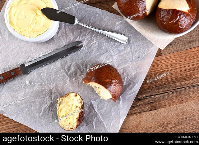 HIgh angle view of a pretzel bun broken in half and buttered. A crock of butter, knife and bowl of whole buns with a broken roll on parchment paper