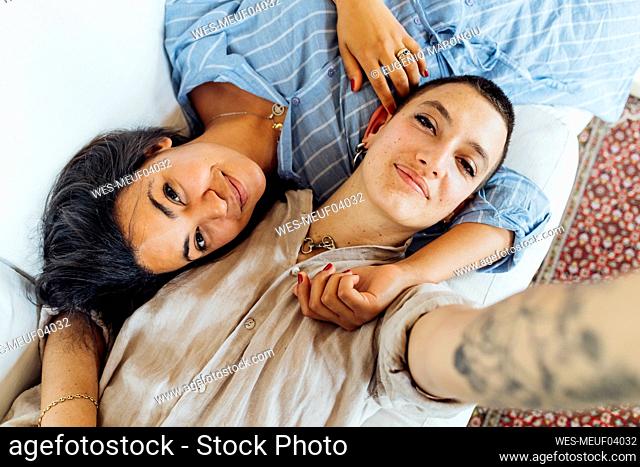 Smiling lesbian couple taking selfie while lying on sofa at home