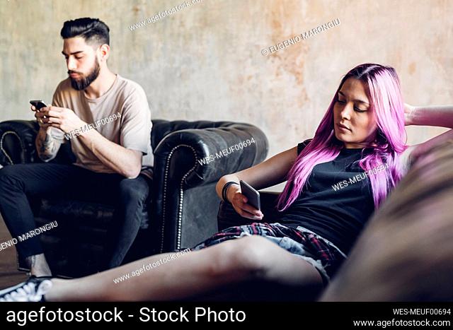 Young man and woman sitting on sofa and armchair in a loft using smartphones
