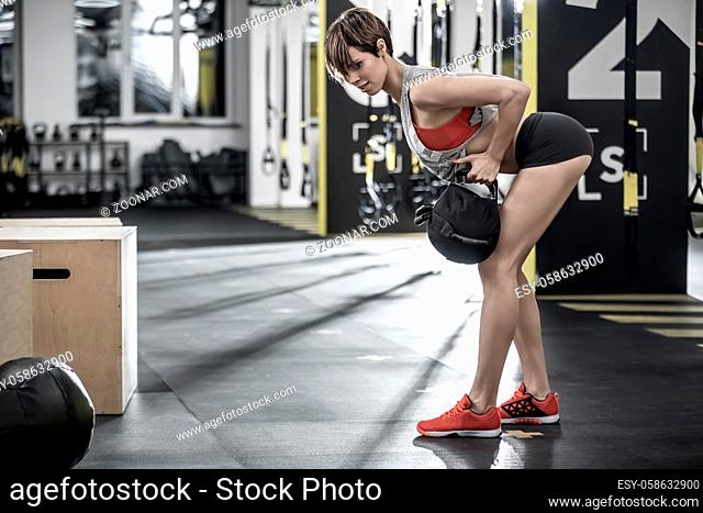 Gorgeous girl with a load does a forward tilt in the gym. She wears a black shorts, red top and sneakers, gray sleeveless. Shoot from the side