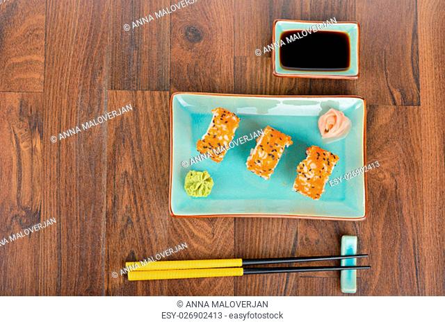 Sushi rolls with masago, served on turquoise plate with pickled ginger, soy sauce and chopsticks on wooden table. Overhead view