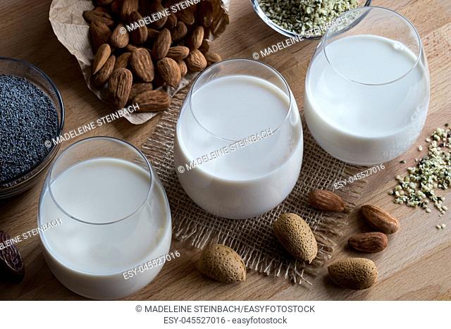 Three glasses of vegan plant milk - almond milk, poppy seed milk and hemp seed milk, with shelled and unshelled almonds, poppy seeds and hemp seeds on a wooden...