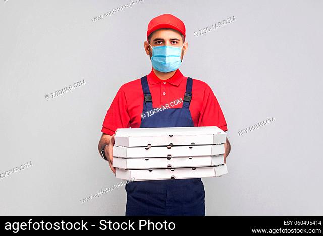 Delivery on quarantine. Young man with surgical medical mask in blue uniform and red t-shirt standing and holding stack of cardboard pizza boxes on grey...