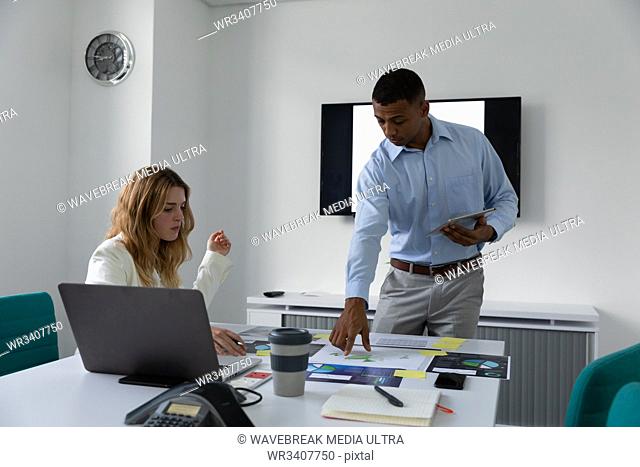 Front view of a young African American man standing holding a tablet computer and talking with a young Caucasian woman sitting at a desk