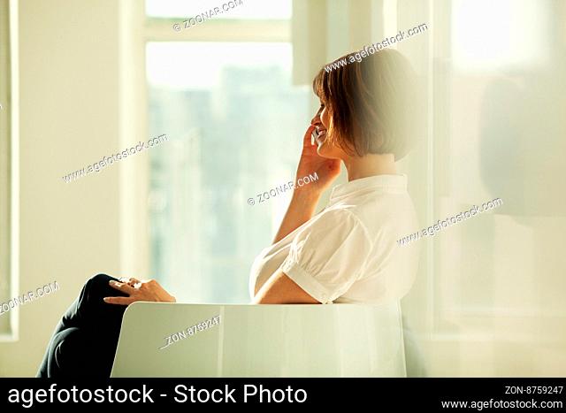 Smiling adult businesswoman talking on smartphone while sitting in chair