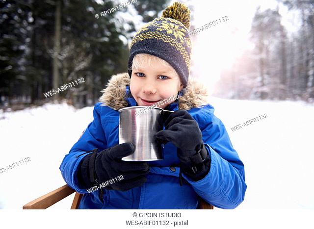 Portrait of smiling little boy sitting on sledge in winter forest drinking tea