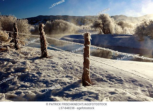 barb wire fence in winter landscape with hoarfrost, steeming river, brilliant sunshine and blue sky, Utah, USA