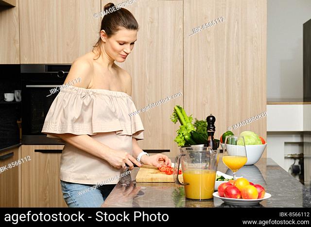 Pregnant woman cutting fresh tomato, making vegetable salad in the kitchen