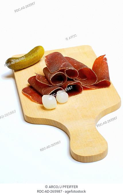 Bundner beef jerky on wooden board, with gherkin and pearl onions / Bündner Trockenfleisch