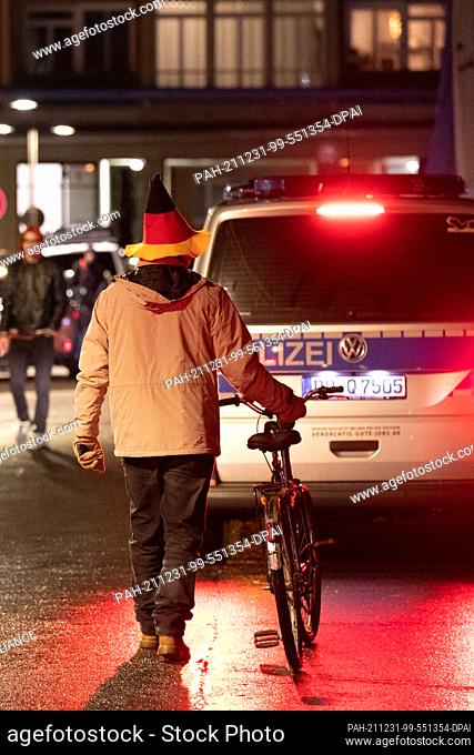 30 December 2021, Saxony, Dresden: A man wearing a cone hat in the colors of the Federal Republic of Germany (black-red-gold) walks on the street pushing his...