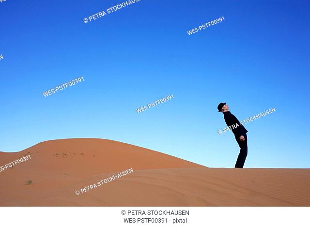 Morocco, Merzouga, Erg Chebbi, man wearing a bowler hat standing crooked on desert dune
