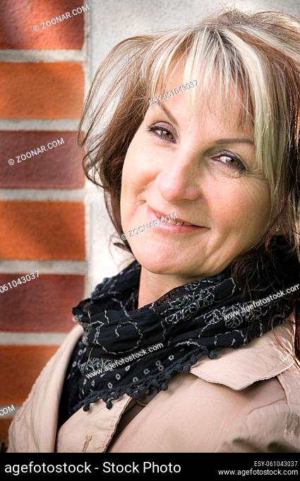 Side close-up of a middle-aged woman leaning her back against an outside wall and looking at the camera with her head tilted