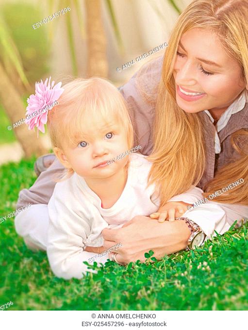 Cheerful young mother playing with her adorable little daughter in the park, lying down on fresh green grass field, happy family life