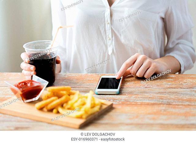 fast food, people, technology and diet concept - close up of woman with smartphone drinking coca cola and eating french fries