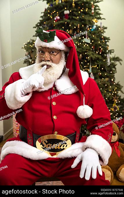 A Black Santa Claus looking into camera and making a shushing gesture