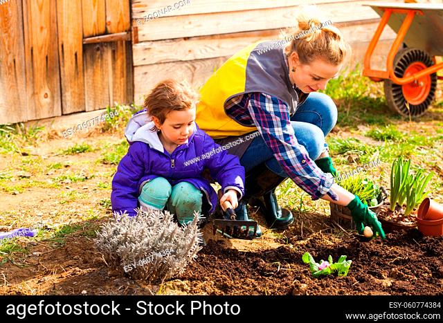 Young attractive blond woman in special costume, working in garden. Adorable preschool girl helping her mother with small rake