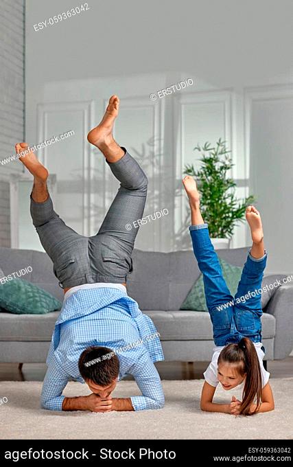 father copy imitate active child girl doing gymnastic handstand exercise at home, dad and daughter stand on hands, sporty family