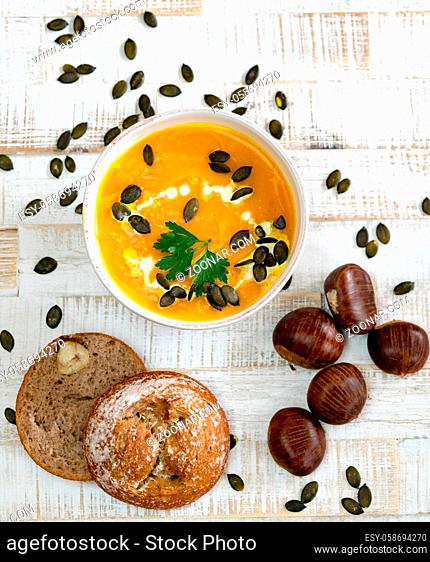 Top down view of organic pumpkin soup in a white bowl framed by chestnuts and chestnut bread and pumpkin seeds