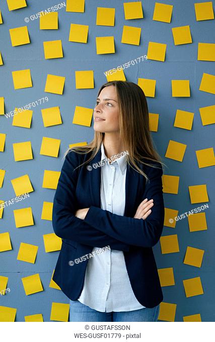 Yong businesswoman standing in front of wall, full of yellow sticky notes, with arms crossed