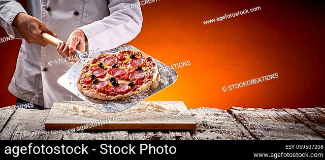 Chef with a delicious homemade pepperoni pizza displaying it on a metal paddle after removing it from the oven in a pizzeria in a rustic panorama banner with...