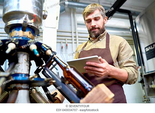 Portrait of middle-aged technician with bushy beard studying operation manual with help of digital tablet while standing at beer bottling equipment