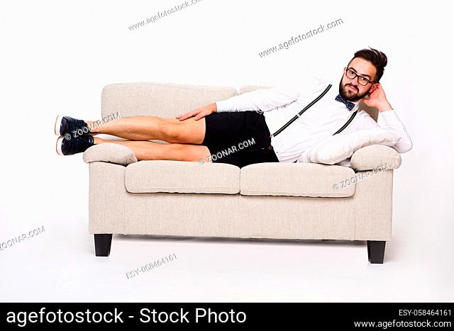Picture of happy smiling handsome hipster man lying on couch or sofa in studio. Bearded man in shirt and shorts looking at camera