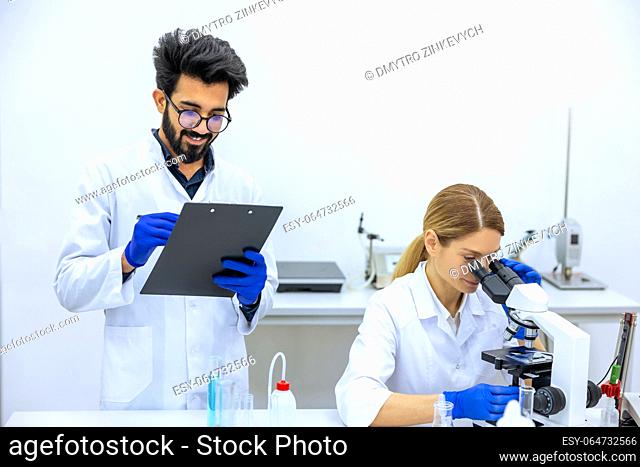 Woman and man scientists in lab coat making notes after doing sample test in laboratory, professional worker analyzes received data together