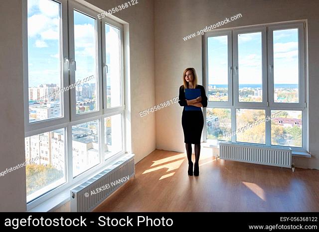 The girl with the folder stands in the middle of a large spacious room with large windows