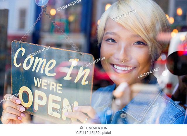 Portrait confident young female shop owner holding Open sign at window