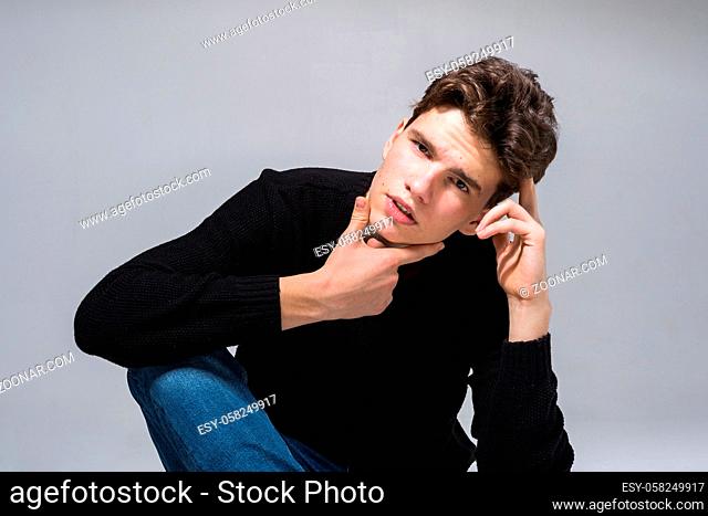 Stylish guy posing in studio sitting on the floor in denim pants and black sweatshirt. Male student portrait full on white background