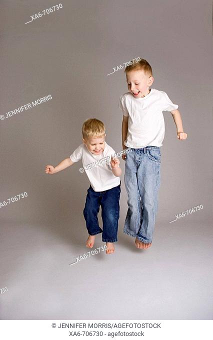 Two blond haired little boyes, aged 6 and 3 years, wearing white t-shirts and blue jeans, smiling and jumping. Studio lighting, grey background