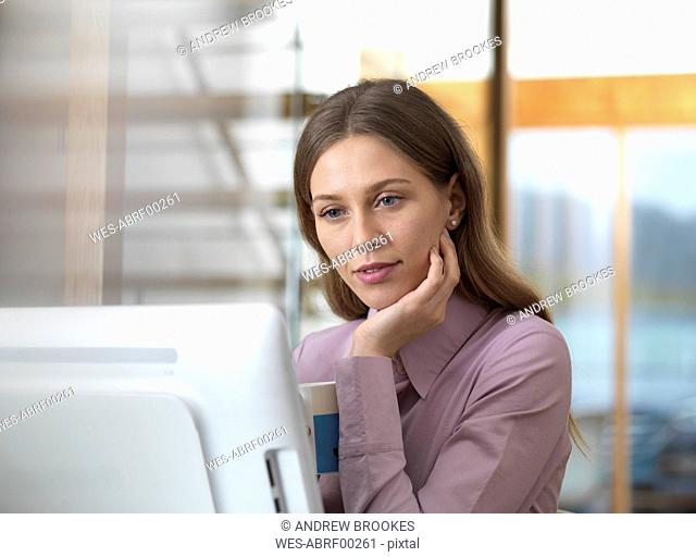 Businesswoman in office holding cup looking at PC