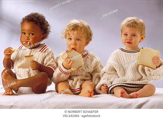 portrait, full-figure, indoor, group of 3 1-year-old children, 1 black and 2 blond boys wearing white pillover sit with a slice of bread in their hands on a...