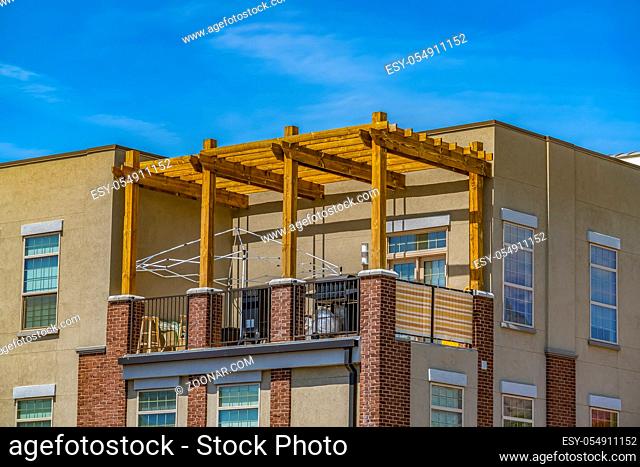Exterior of a residential building illuminated by sunlight against blue sky. A wooden pergola is built on the open balcony of this building