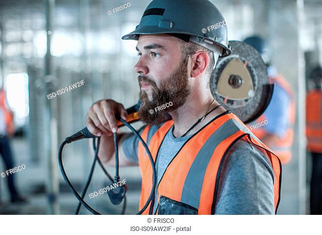 Young male builder carrying circular saw on shoulder on construction site