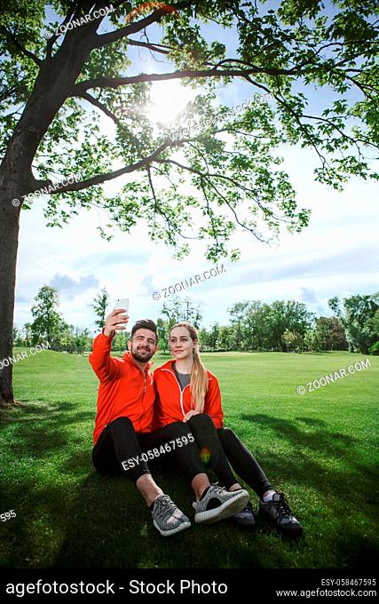 Sporty couple making selfies in green park or forest. Man and woman sittin on green grass and posing for photo camera