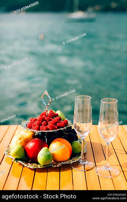 Two-storeyed plate with fruits: raspberries, peach, apples, figs, pears, grapes. And two glasses for champagne on the table near the sea