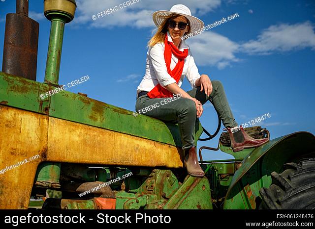 Urban woman in casual clothing sitting on an old tractor against the blue sky in the farmland