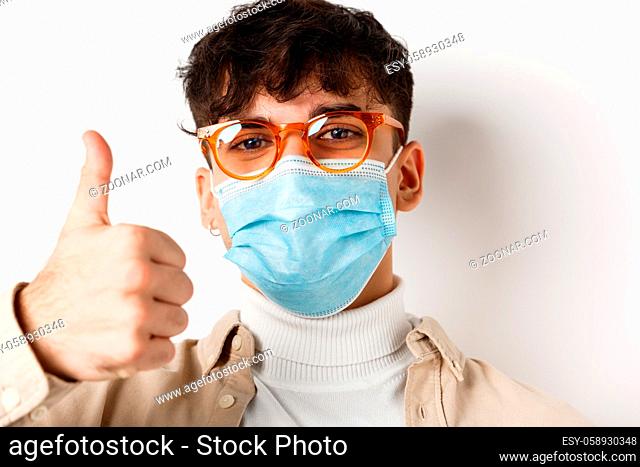 Portrait of young man in glasses and medical mask showing thumb up, smiling with eyes, standing on white background. Covid-19 and pandemic concept