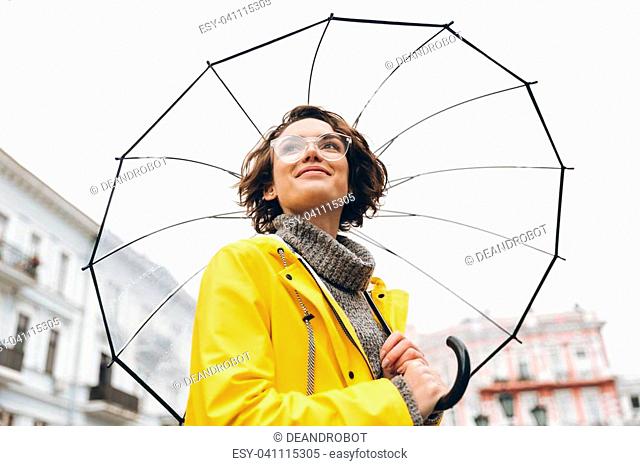 View from beneath of positive woman in yellow raincoat and glasses standing in street under big transparent umbrella, during grey rainy day