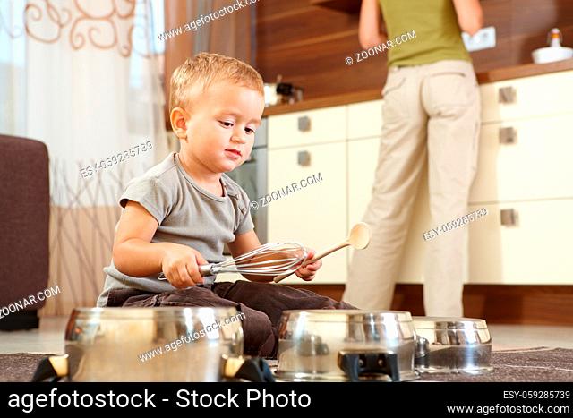 Little boy sitting on carpet in kitchen playing with cooking pots, mother preparing food in background