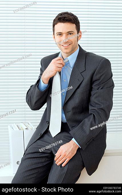 Businessman wearing grey suit and blue shirt, sitting in office, adjusting his tie, smiling