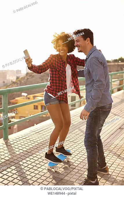 Curly haired girl posing on a skateboard while boy holds her to take a selfie of them with big toothy smiles