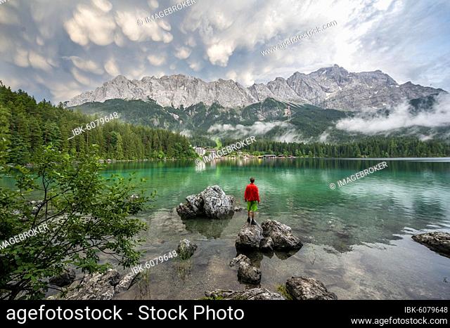 Young man standing on a rock on the shore, Eibsee lake in front of Zugspitze massif with Zugspitze, dramatic Mammaten clouds, Wetterstein range, near Grainau