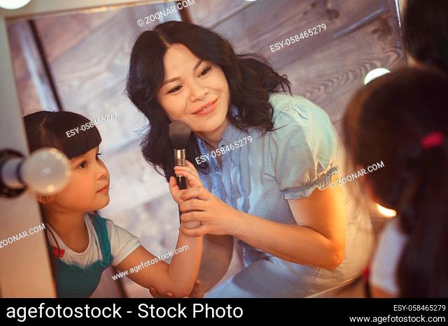 Picture of mother and daughter doing makeup while looking at mirror. Little lady using brush to do makeup for her mother